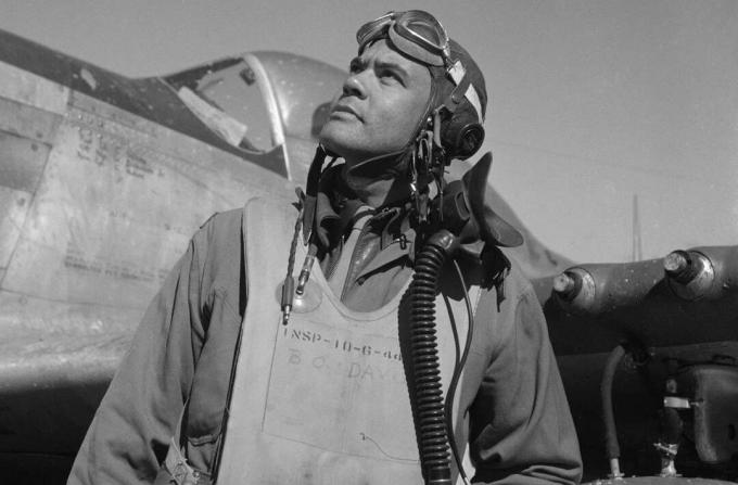 Бенджамин О. Дейвис в полетен костюм и шлем, стоящ пред изтребител P-51 Mustang.